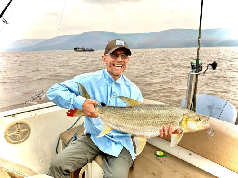 A smiling fisherman proudly displays a large, silvery fish he just caught while on a boat on a cloudy day.