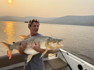 A man proudly holds a large tigerfish he caught on a boat at sunset, with a hazy landscape in the background.