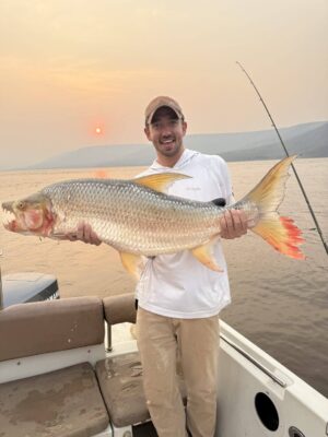A smiling man in a cap and white shirt holds a large, silver fish with orange fins while standing on a boat at sunset.